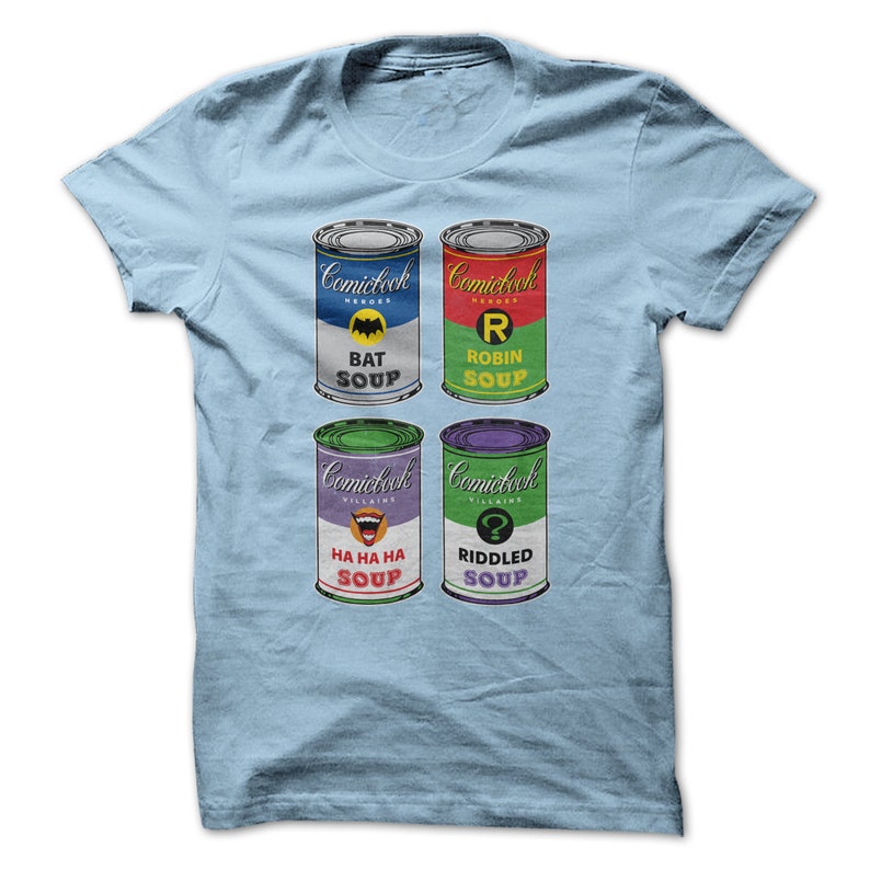 May include: A light blue t-shirt with a graphic of four soup cans featuring Batman, Robin, The Joker, and The Riddler. The cans are labeled "Comiclock Heroes" and "Comiclock Villains".