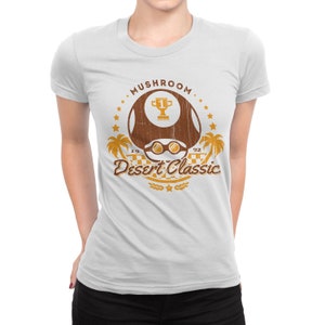 May include: White t-shirt featuring a brown mushroom graphic with the text "Mushroom Desert Classic 1992". The design includes a trophy, sunglasses, palm trees, and stars. The shirt is a crew neck style.