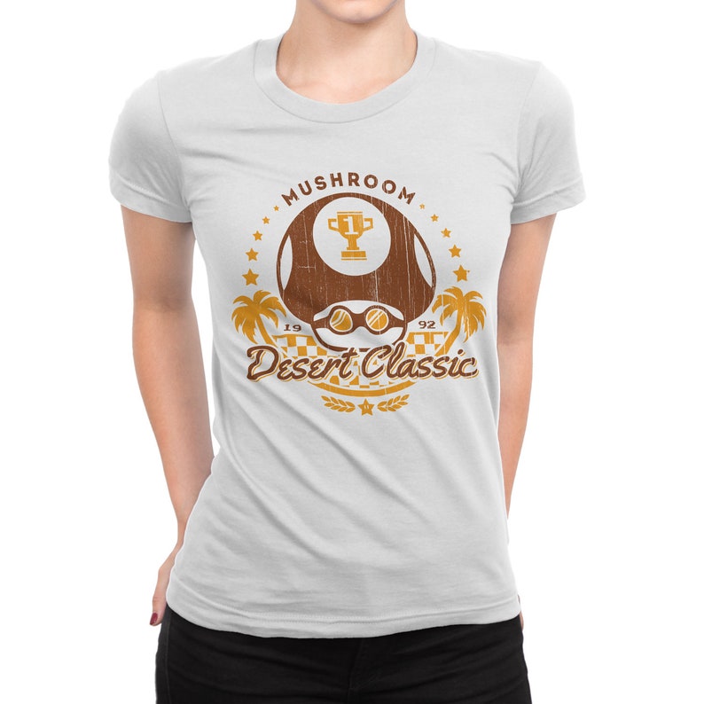 May include: White t-shirt featuring a brown mushroom graphic with the text "Mushroom Desert Classic 1992". The design includes a trophy, sunglasses, palm trees, and stars. The shirt is a crew neck style.