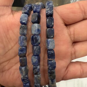 May include: Three strands of blue sodalite gemstone beads. The beads are square-shaped with a mix of dark and light blue hues. The beads are strung together, creating a bracelet or necklace.