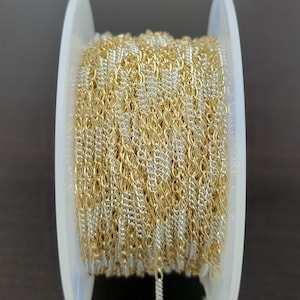 May include: A spool of gold and silver-colored chain. The chain is tightly wound around a white plastic spool. The chain has a delicate, interwoven design, with alternating gold and silver links. The chain is likely used for jewelry making or other craft projects.