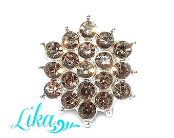 May include: A silver-toned, snowflake-shaped brooch with multiple brown-colored rhinestones. The brooch has a central cluster of rhinestones surrounded by a ring of smaller stones. The word "Lika" is written in a teal cursive font.
