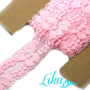 May include: A roll of delicate pink lace ribbon with a floral pattern. The lace is displayed on a brown cardboard spool. The image is well-lit, showcasing the intricate details of the lace. The ribbon is ideal for sewing and craft projects.