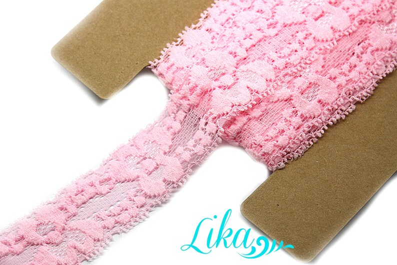 May include: A roll of delicate pink lace ribbon with a floral pattern. The lace is displayed on a brown cardboard spool. The image is well-lit, showcasing the intricate details of the lace. The ribbon is ideal for sewing and craft projects.