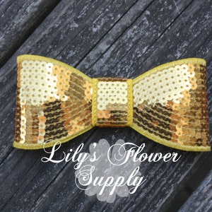 Op de afbeelding: Een gouden strik met pailletten en een gele bies. De strik ligt op een donker houten achtergrond. "Lily's Flower Supply" staat onder de strik.