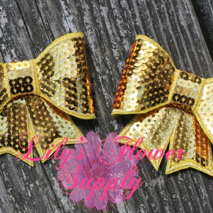 May include: Two gold sequin bows with yellow trim. The bows are on a wooden surface.