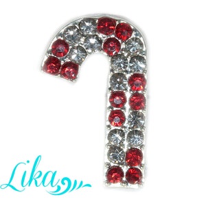 May include: A candy cane-shaped brooch, encrusted with red and clear rhinestones, set in a silver-tone metal. The design alternates red and clear stones, creating a festive holiday accessory. The word "Lika" is written in turquoise script.