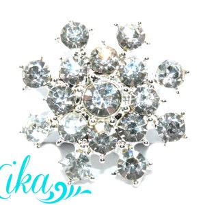 May include: A silver-toned, flower-shaped brooch with numerous clear, sparkling rhinestones. The brooch has a central cluster of stones surrounded by radiating petals, each with a single stone. The word "Lika" is written in turquoise script.