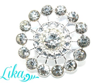 May include: A silver-toned brooch with a circular design. The brooch features a large, central clear rhinestone surrounded by smaller rhinestones, creating a sparkling, floral-like pattern. The word "Lika" is written in cursive in the bottom left corner.