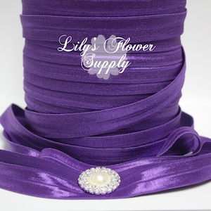 May include: A roll of purple elastic ribbon with a single strand of ribbon laying on top of the roll. The ribbon has a small, clear rhinestone embellishment with a pearl center.