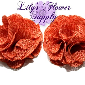 May include: Two orange-red fabric flower embellishments with a textured, woven appearance. The flowers are in full bloom, with multiple layers of petals. The text "Lily's Flower Supply" is visible in the upper portion of the image.