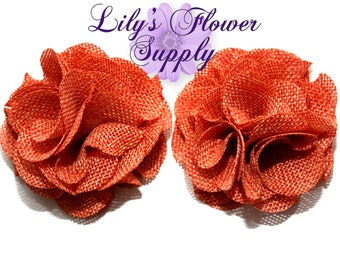 May include: Two orange-red fabric flower embellishments with a textured, woven appearance. The flowers are in full bloom, with multiple layers of petals. The text "Lily's Flower Supply" is visible in the upper portion of the image.