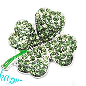 May include: A silver-toned shamrock brooch with green rhinestone accents. The three-leaf clover design has a green enamel stem and is set against a white background. The word "Lika" is written in a teal script.