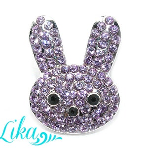 Può includere: Spilla a forma di coniglietto color argento, incastonata con numerosi strass viola chiaro. Il coniglietto ha occhi neri e orecchie dritte. La parola "Lika" è scritta in turchese in basso. La spilla è su sfondo bianco.
