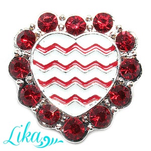 May include: A heart-shaped jewelry piece with a silver-colored metal frame. The center features a white background with red zigzag lines. The heart is surrounded by a circle of red gemstones. The word "Lika" is written in a teal script font.