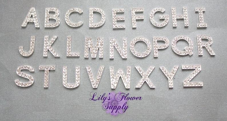 Puede incluir: Un conjunto de 26 letras plateadas, cada una decorada con diamantes de imitaci&oacute;n transparentes, que deletrean el alfabeto de la A a la Z. Las letras est&aacute;n dispuestas en tres filas sobre un fondo texturizado gris claro. Las letras miden aproximadamente 2,5 cm de alto.