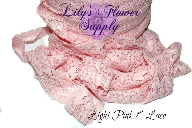 May include: Light pink lace ribbon, 1 inch wide, is displayed in a rolled form. The delicate fabric is intricately patterned with floral designs. The text "Lily's Flower Supply" is visible in the image.