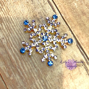 May include: A gold snowflake embellishment with blue and clear rhinestones. The snowflake is on a wood background. Lily's Flower Supply.