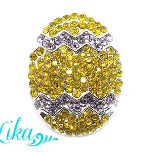 May include: An Easter egg-shaped brooch with a silver-coloured metal base. The egg is covered in yellow and purple rhinestones, arranged in a pattern. The brooch has a shiny, reflective surface.