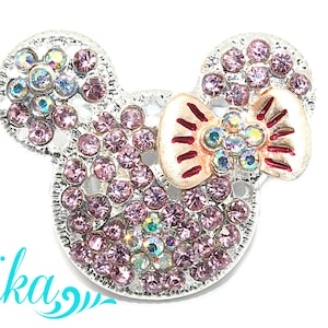 May include: A silver-toned, rhinestone-encrusted Mickey Mouse head with a pink and red bow. The bow has a metallic finish.