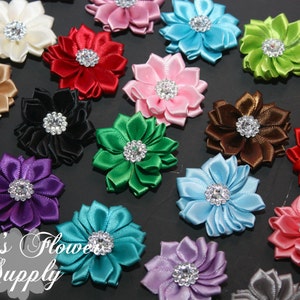 Op de afbeelding: Een verzameling kleurrijke satijnen bloemversieringen met strass-middens. De bloemen zijn in verschillende kleuren, waaronder wit, goud, rood, zwart, groen, roze, bruin, blauw, paars en turquoise. De tekst "Lily's Flower Supply" is zichtbaar.