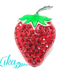 Puede incluir: Broche con forma de fresa con cuerpo rojo incrustado de joyas, hoja verde y tallo plateado. El broche está sobre un fondo blanco. La palabra "Lika" está escrita en turquesa.