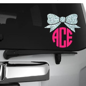May include: A silver glitter bow with a pink circle containing the letters "ACE" in white. The bow is on a silver car with a black rear window. There are two other examples of the bow with different letters and colors.