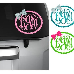 May include: A pink car decal featuring a monogram within a beaded circle, topped with a glittery bow. The decal is displayed on a car's rear window. Additional decals in teal and green with bows are also shown.