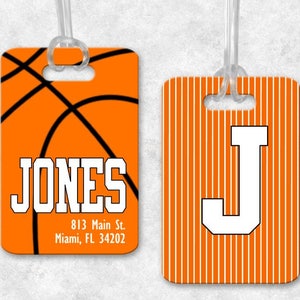 May include: Two orange luggage tags with white text. The first tag features a basketball design and the text "JONES 813 Main St. Miami, FL 34202". The second tag has white stripes and the letter "J" in white.