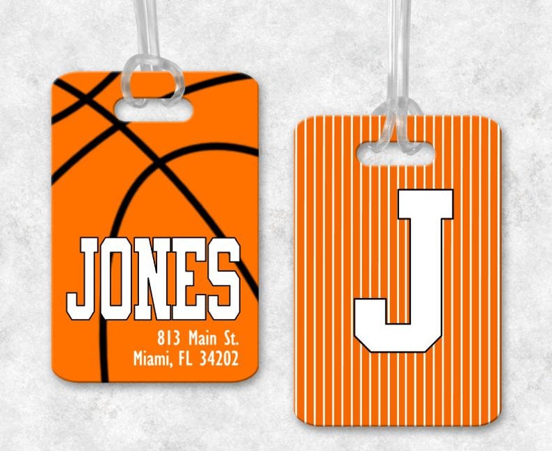 May include: Two orange luggage tags with white text. The first tag features a basketball design and the text "JONES 813 Main St. Miami, FL 34202". The second tag has white stripes and the letter "J" in white.