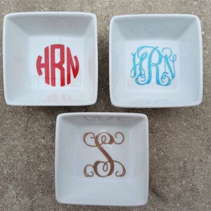 May include: Three white ceramic ring dishes with different monogram designs. The first dish has a red monogram "HRN", the second dish has a blue monogram "HRN", and the third dish has a gold monogram "S".