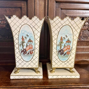 Puede incluir: Un par de jarrones de cerámica blanca con detalles dorados y escenas pintadas de inspiración asiática. Los jarrones tienen una base cuadrada con cuatro patas doradas y una parte superior festoneada.