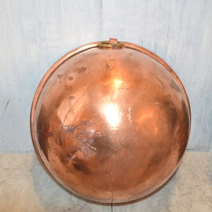 May include: A large, round copper bowl with a brass handle. The bowl is shiny and has a few scratches on the surface.