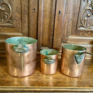 Puede incluir: Tres ollas de cobre de diferentes tamaños con asas de metal oscuro. Las ollas están sobre una superficie de madera.