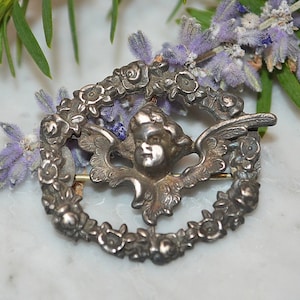 May include: Silver tone brooch featuring a cherub with wings surrounded by a wreath of roses.