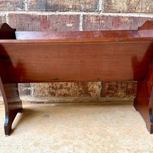 Op de afbeelding: Een houten poppenwieg met een donkerbruine afwerking. De wieg heeft een rechthoekige vorm met afgeronde hoeken en staat op vier poten.