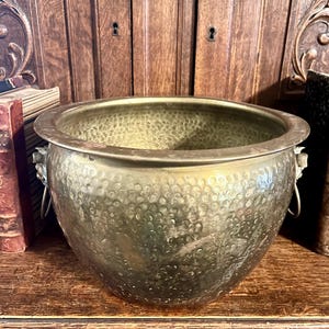 May include: A hammered brass planter with a wide, round bowl shape. The planter has two small handles and a textured surface. The background includes a wooden cabinet and books, suggesting an antique or vintage aesthetic.