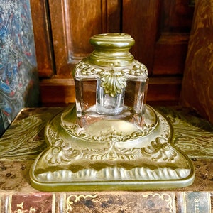 May include: A vintage brass inkwell with a glass jar and ornate details. The inkwell is sitting on top of a leather-bound book.
