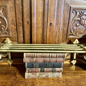 May include: A brass towel rack with three bars, mounted on a wooden surface. The rack is holding a stack of antique books with brown and black covers. The background features carved wooden panels.