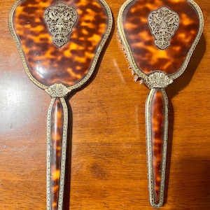 Op de afbeelding: Een handspiegel en haarborstelset met een bruin en oranje schildpadpatroon. Beide items hebben een sierlijk zilverkleurig metalen frame en een decoratief filigraan embleem in het midden van de achterkant met een lange steel.