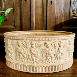 Puede incluir: Una jardinera beige, de forma ovalada, con un relieve detallado de querubines y vides. La jardinera tiene una base acanalada y un borde liso. El fondo es una superficie de madera oscura y un libro antiguo.