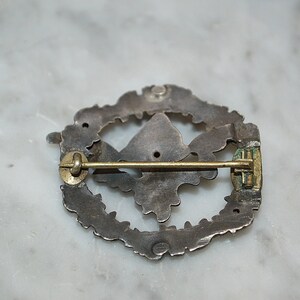May include: The back of a silver pin with a detailed design. The pin has a gold-colored clasp and a pin back.