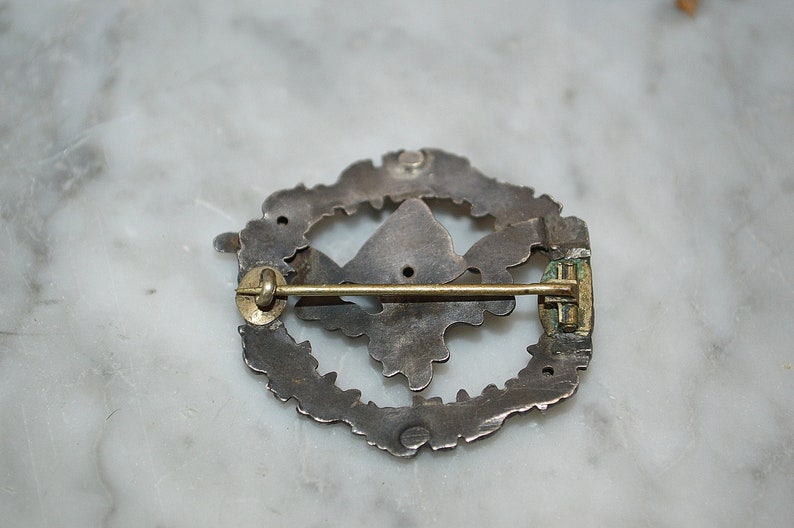 May include: The back of a silver pin with a detailed design. The pin has a gold-colored clasp and a pin back.