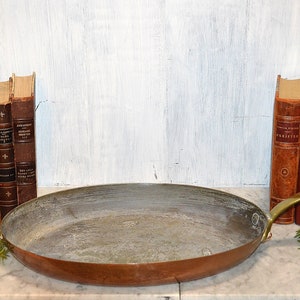 May include: A vintage copper oval pan with a brass handle. The pan is sitting on a marble surface with a stack of antique books on either side.