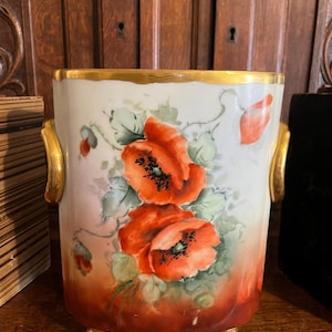May include: A cylindrical porcelain planter featuring hand-painted red poppy flowers with green foliage on a white and gradient orange background. The vessel has gold-painted handles on the sides, a gold rim, and stands on three small gold feet.