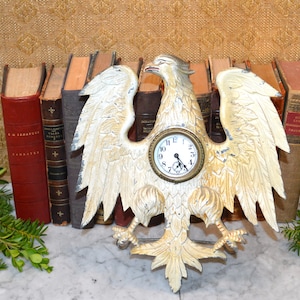 May include: A white and gold painted eagle clock with a round clock face. The clock is sitting on a shelf with a row of books behind it.