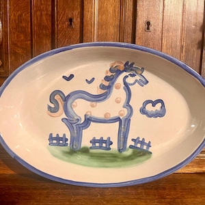 May include: An oval ceramic platter with a white base and a blue rim. The center features a hand-painted blue horse with light pink spots, standing on a green patch of grass behind a blue fence. Two blue birds and a blue cloud are painted above.