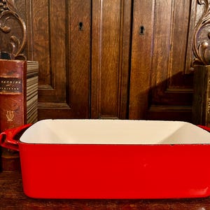 May include: A rectangular red enamel baking dish with a white interior. The dish has two handles and is set against a backdrop of antique books and a wooden cabinet.