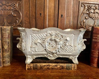 May include: A weathered, rectangular, cast-iron planter with ornate floral and scrollwork details. The planter is painted in a distressed, light gray finish and has four small feet. It is displayed on a wooden surface with antique books in the background.