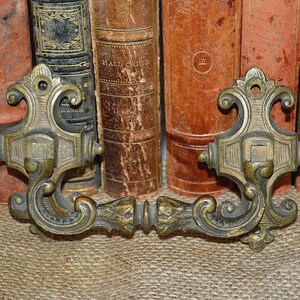 Pode incluir: Puxador de gaveta antigo de latão com detalhes em rolos e uma placa central quadrada. O puxador está em bom estado com acabamento em pátina.